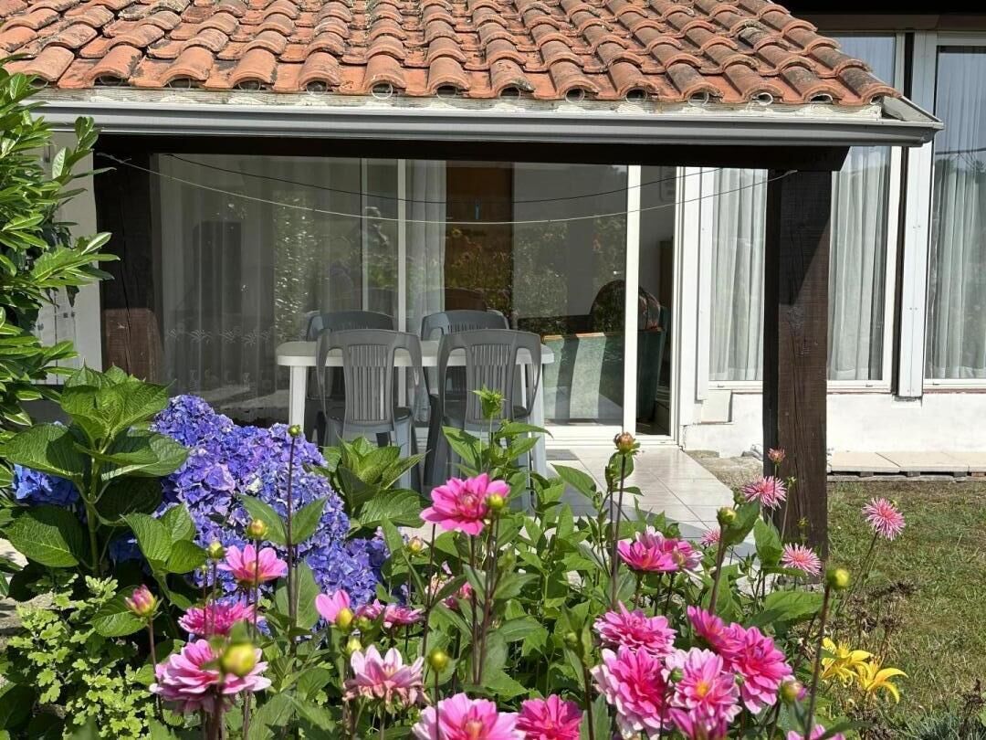 Eine überdachte Terrasse mit Tisch und Stühlen, ein Garten mit verschiedenen Blumen und ein Haus mit Ziegeldach und großen Fenstern.