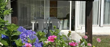 Un patio cubierto con una mesa y sillas, un jardín con diversas flores y una casa con techo de tejas y ventanas amplias.
