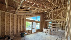 Een kamer in aanbouw met zichtbare houten balken, grote ramen en bouwmaterialen verspreid rondom.