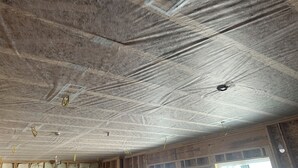 Een kamer met zichtbare plafondisolatie, houten balken en een raam met plastic afdekking.