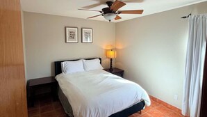 Un dormitorio con cama, ventilador de techo, mesita de noche, lámpara y ventana con cortinas.