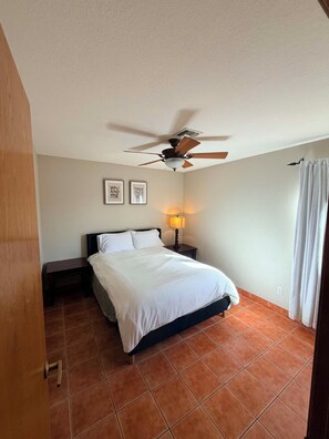 Un dormitorio con cama, ventilador de techo, mesita de noche, lámpara y ventana con cortinas.