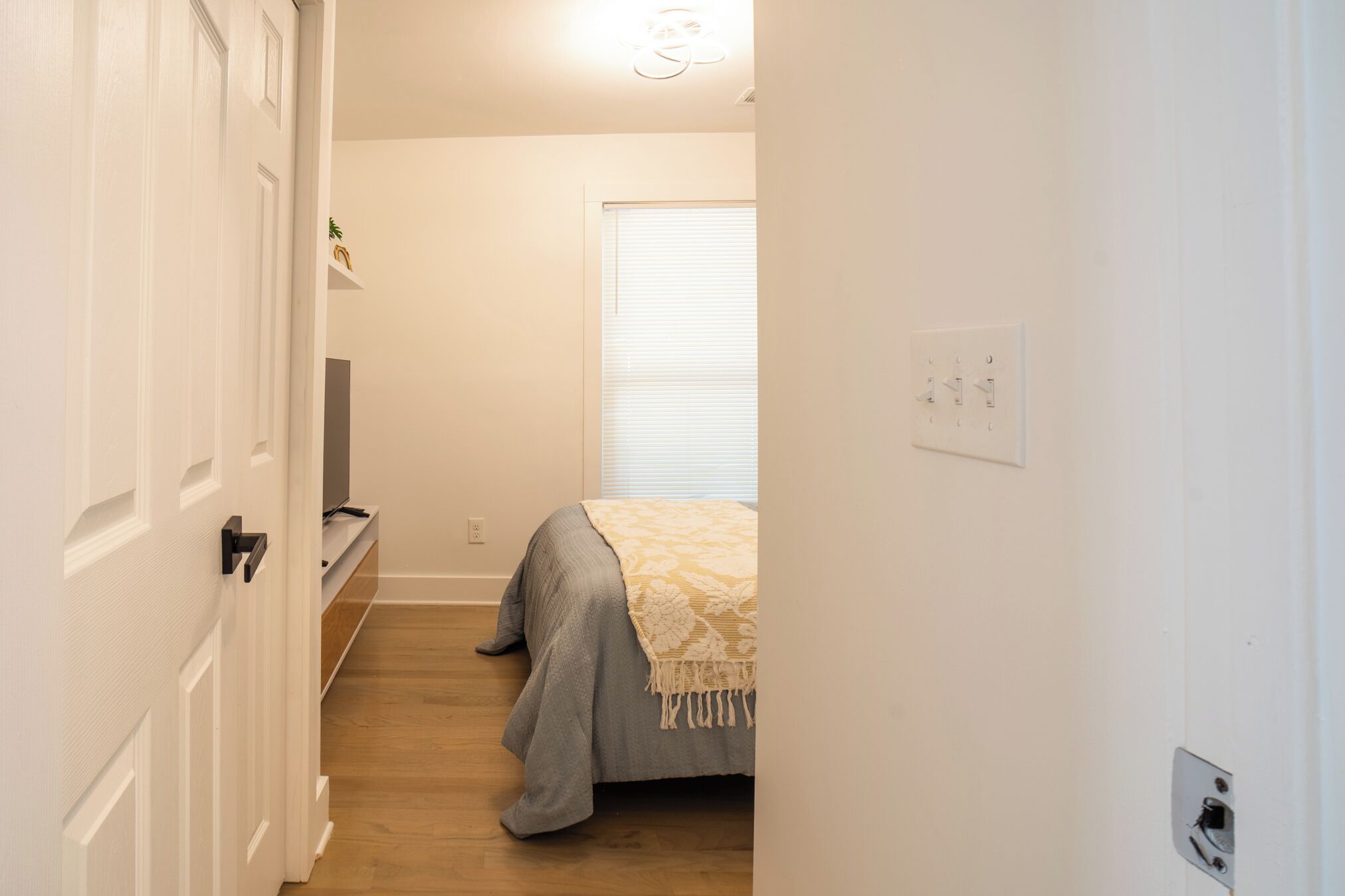 A narrow hallway leading to a bedroom with a bed, a window with blinds, a door, and a wall socket.
