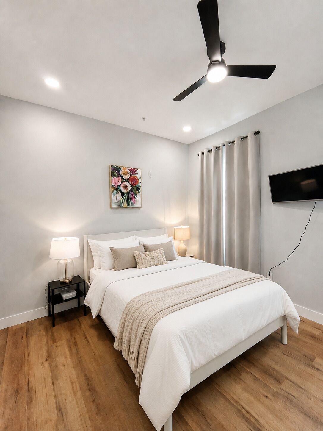 A bedroom with a bed, two bedside tables with lamps, a ceiling fan, a painting, a TV, and a window with curtains.