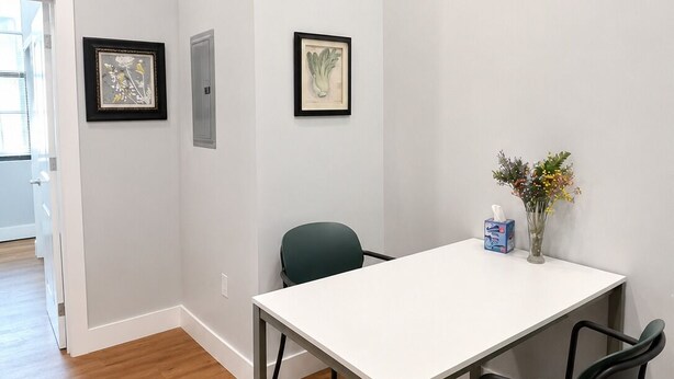 Ein Zimmer mit einem weißen Tisch, zwei Stühlen, einem gerahmten Kunstwerk und einer Vase mit Blumen.