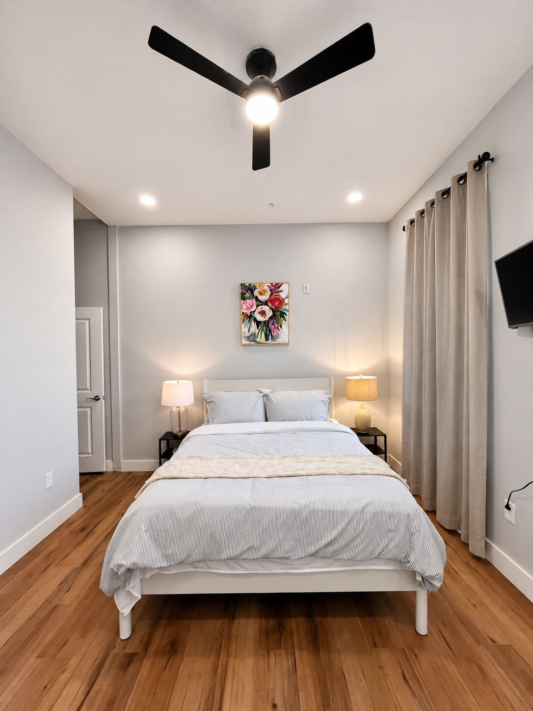 Une chambre à coucher comprenant un lit, deux tables de chevet avec des lampes, un ventilateur de plafond, un téléviseur fixé au mur et une porte.