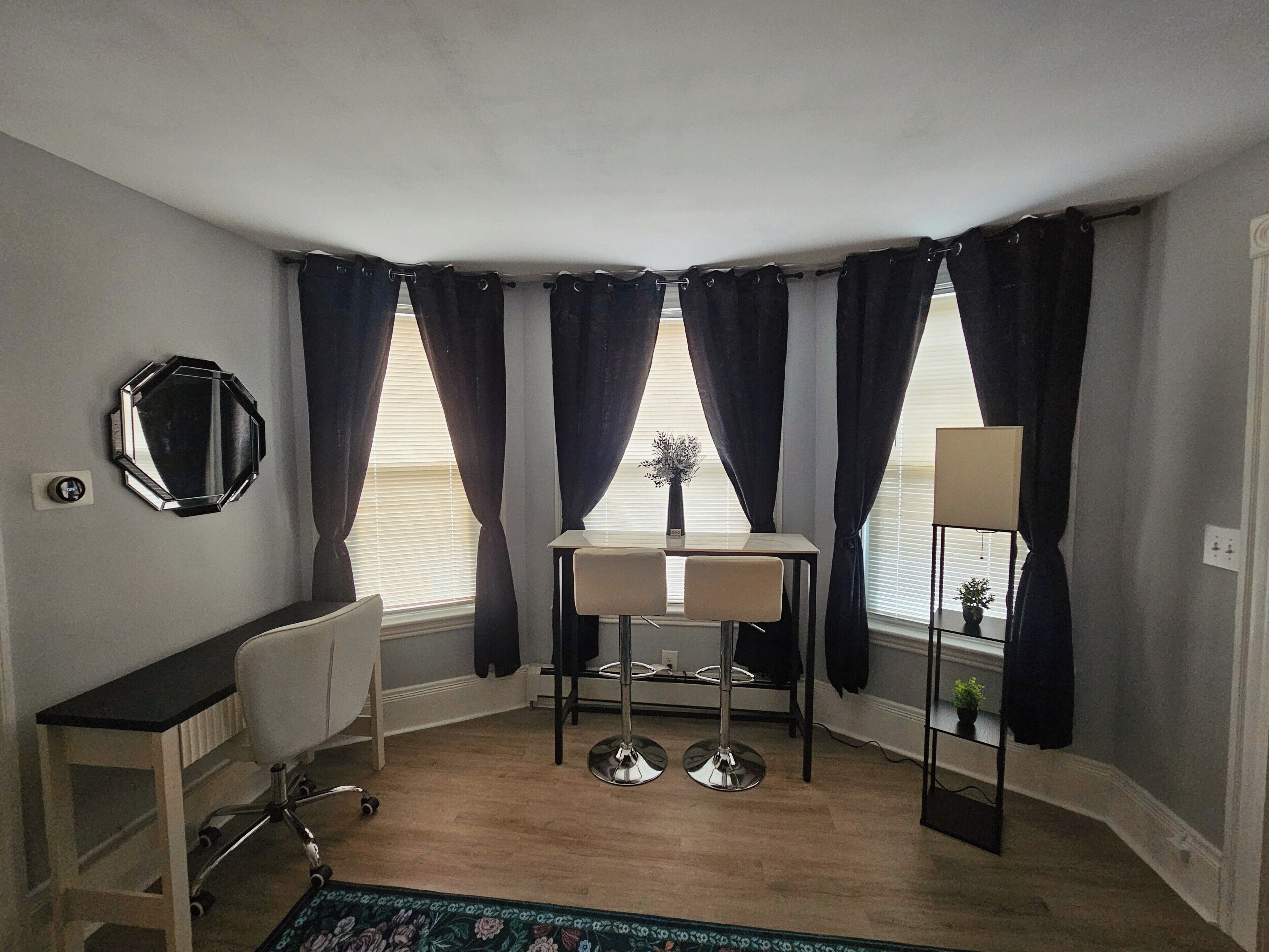 Habitación en esquina con escritorio, silla, espejo y dos ventanas con cortinas.