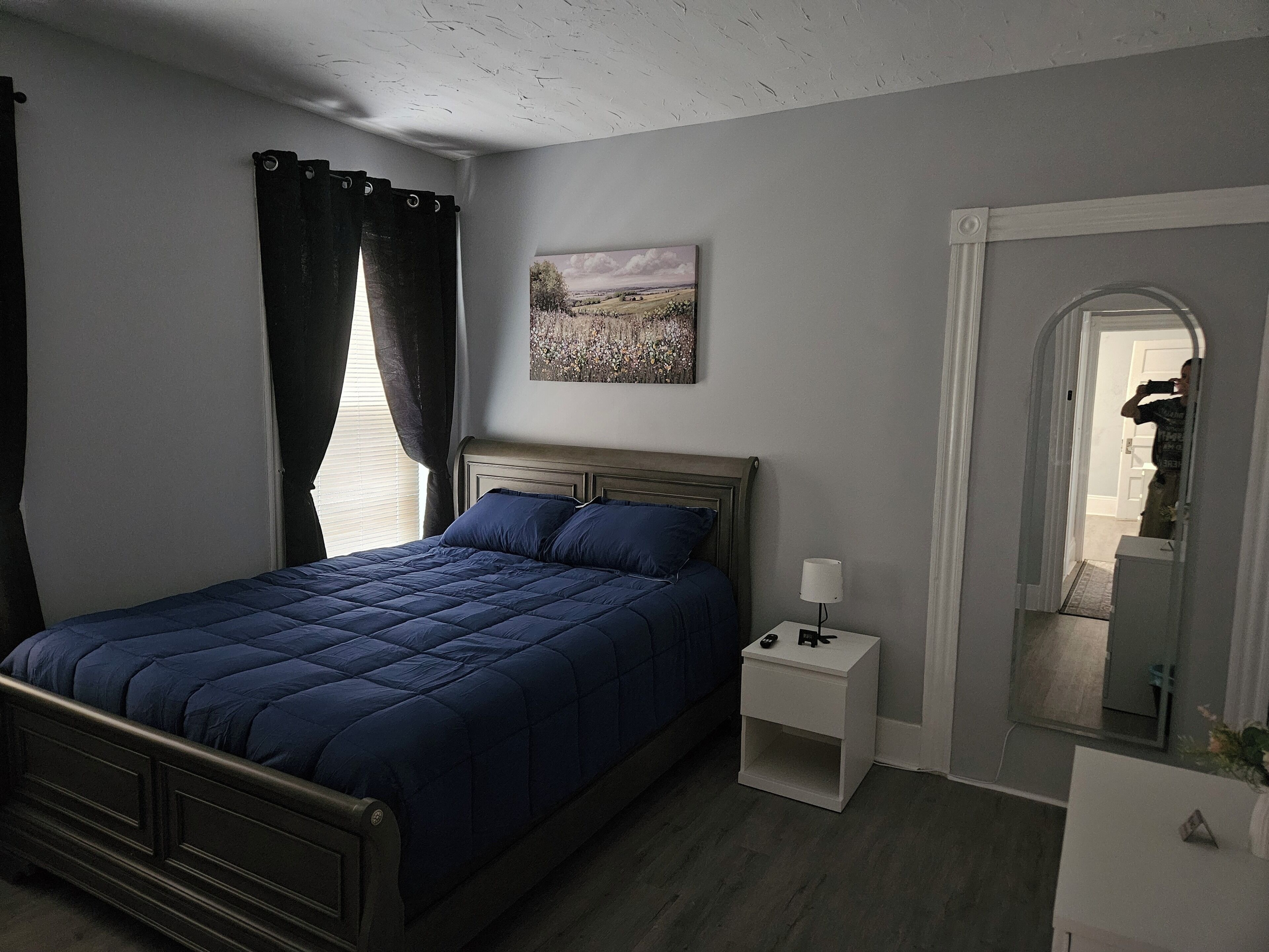A bedroom with a bed, a nightstand, a bedside lamp, a painting on the wall, and a mirror in the corner.