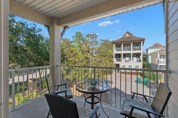 A balcony with two wicker chairs and a small round table, overlooking a street with buildings and trees. - Tahitian Treasure - Villages of Crystal Beach (Destin)