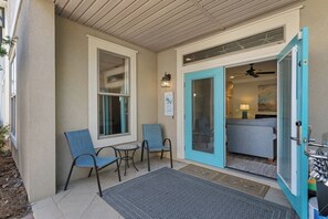 A covered outdoor seating area with chairs and a table, a bedroom with a bed and a ceiling fan, and a blue door leading to the bedroom. - Tahitian Treasure - Villages of Crystal Beach (Destin)