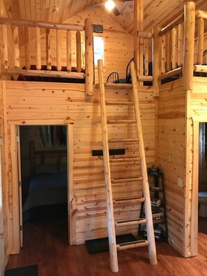 A wooden cabin interior with a ladder leading to a loft area, a door to a bedroom, and a small shelf with items.