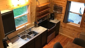 A compact kitchen with a sink, stove, and wooden cabinets. There is a window with a view outside, a table with a tic-tac-toe game, and a shelf with wine glasses.