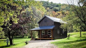 A rustic cabin with a porch, surrounded by lush greenery and a gravel driveway.
