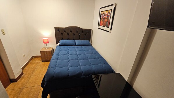 A small, neatly arranged bedroom with a single bed, a bedside table with a lamp, and a framed picture on the wall.