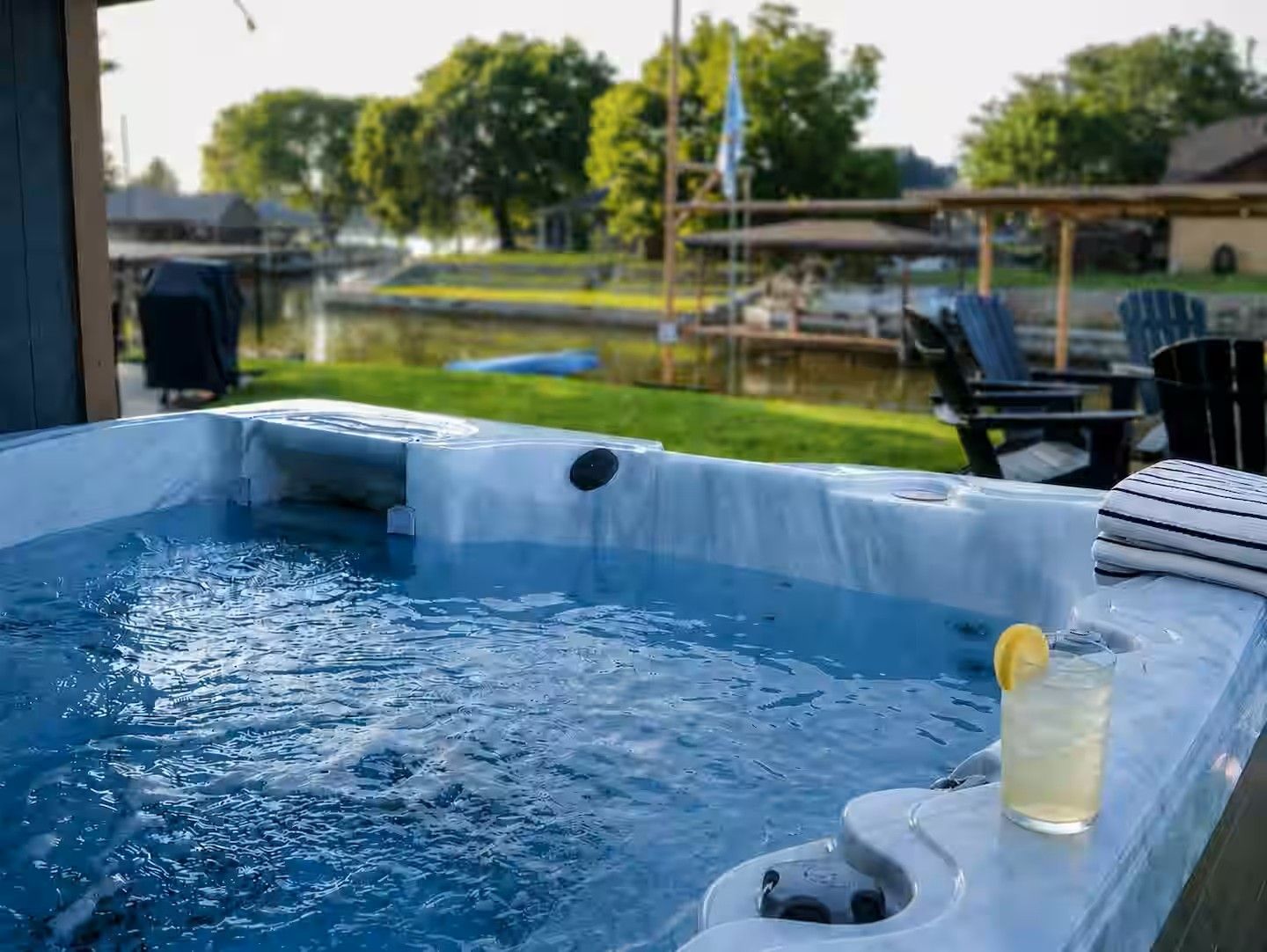 A hot tub with a drink on the side, surrounded by outdoor furniture and a view of a lake.