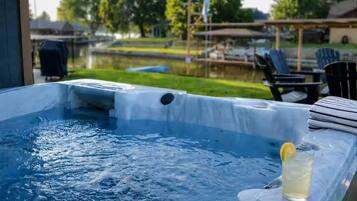 A hot tub with a drink on the side, surrounded by outdoor furniture and a view of a lake.
