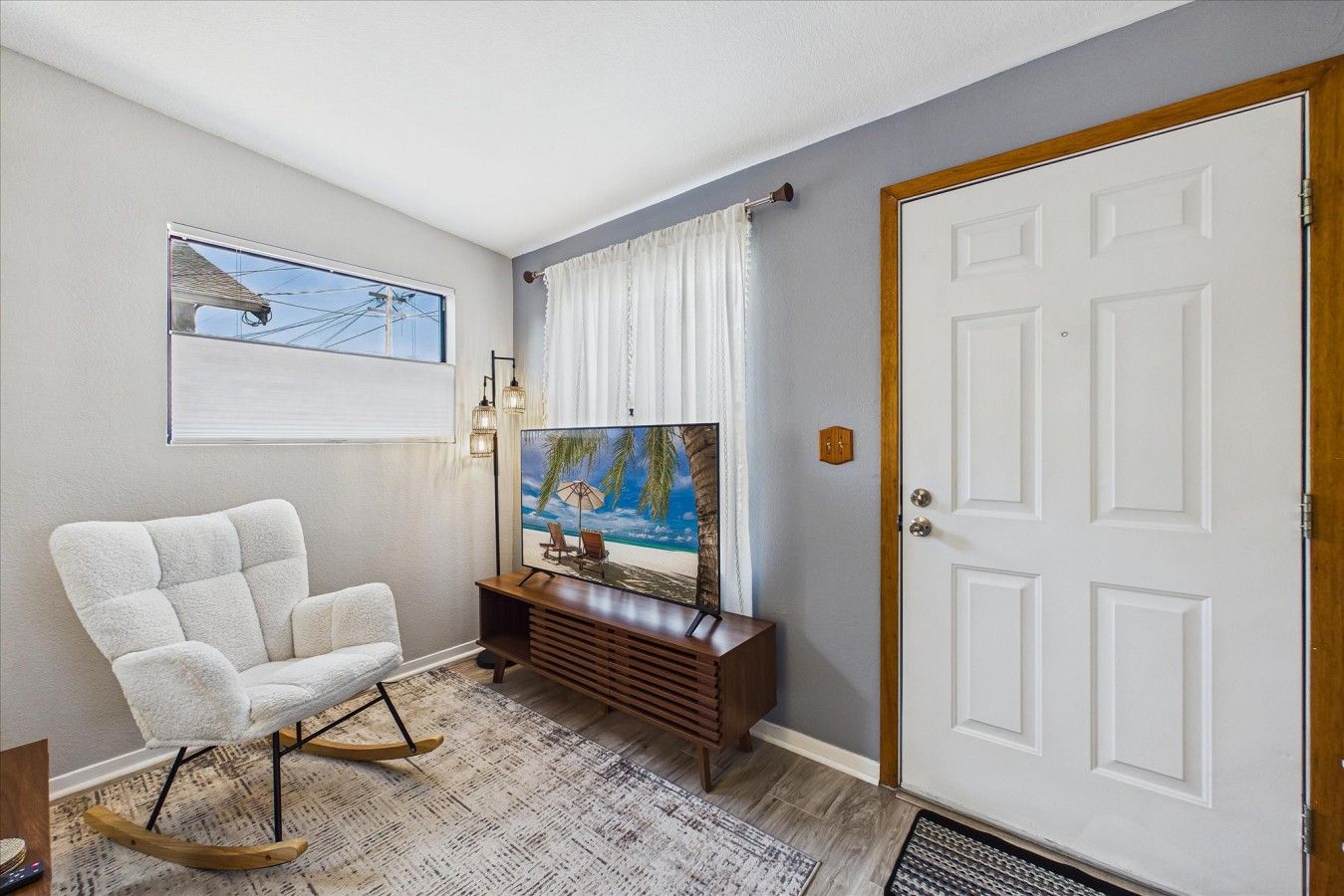 A room with a white armchair, a television on a wooden stand, a door, and a window with a view of the sky.