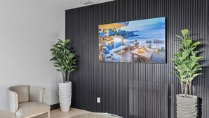 Sala de estar moderna com uma televisão grande fixada em uma parede escura, duas plantas em vasos, um sofá bege e uma mesa de centro de madeira.