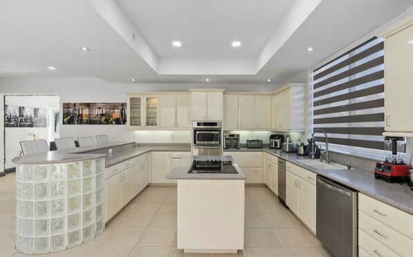A modern kitchen with a central island, built-in appliances, and a large window with blinds.