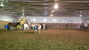 Une carrière d’équitation intérieure avec un sol en terre battue, un plafond en métal et un éclairage suspendu.