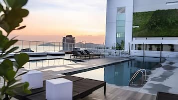 Uma área de piscina no terraço com espreguiçadeiras e guarda-corpo de vidro, com vista para a paisagem urbana ao pôr do sol.
