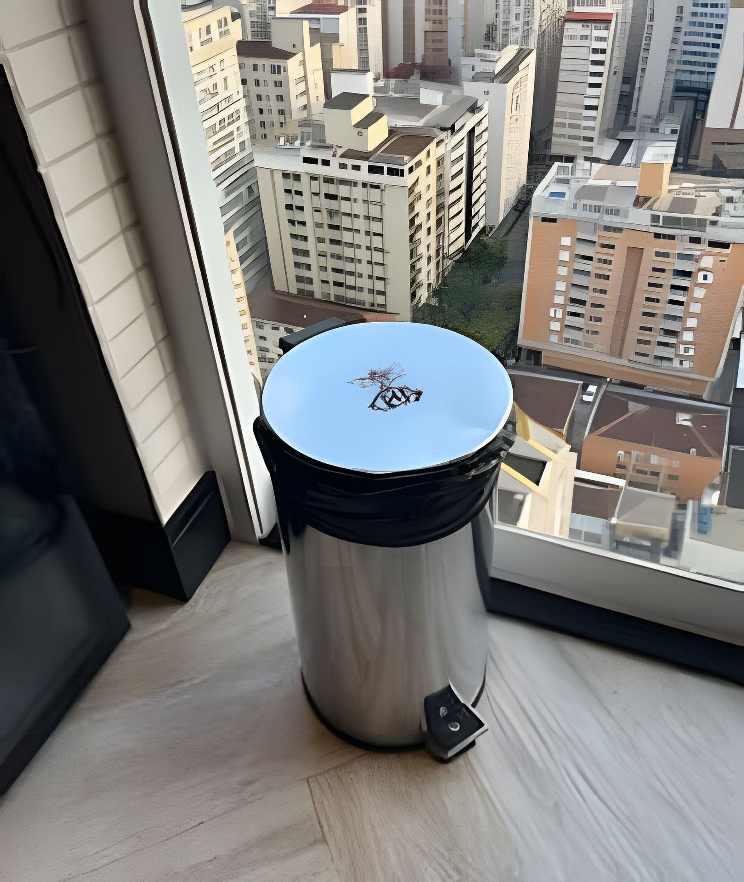 Un cubo de basura metálico con tapa, situado sobre un suelo de madera cerca de una ventana con vistas a un paisaje urbano.