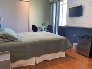 Quarto de hotel com cama, escrivaninha, cadeira, televisão e uma pequena cozinha equipada.
