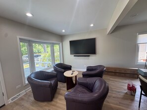 A living room with a flat-screen TV mounted on the wall, leather sofas arranged around a small round table, and a sliding glass door leading to an outdoor area.
