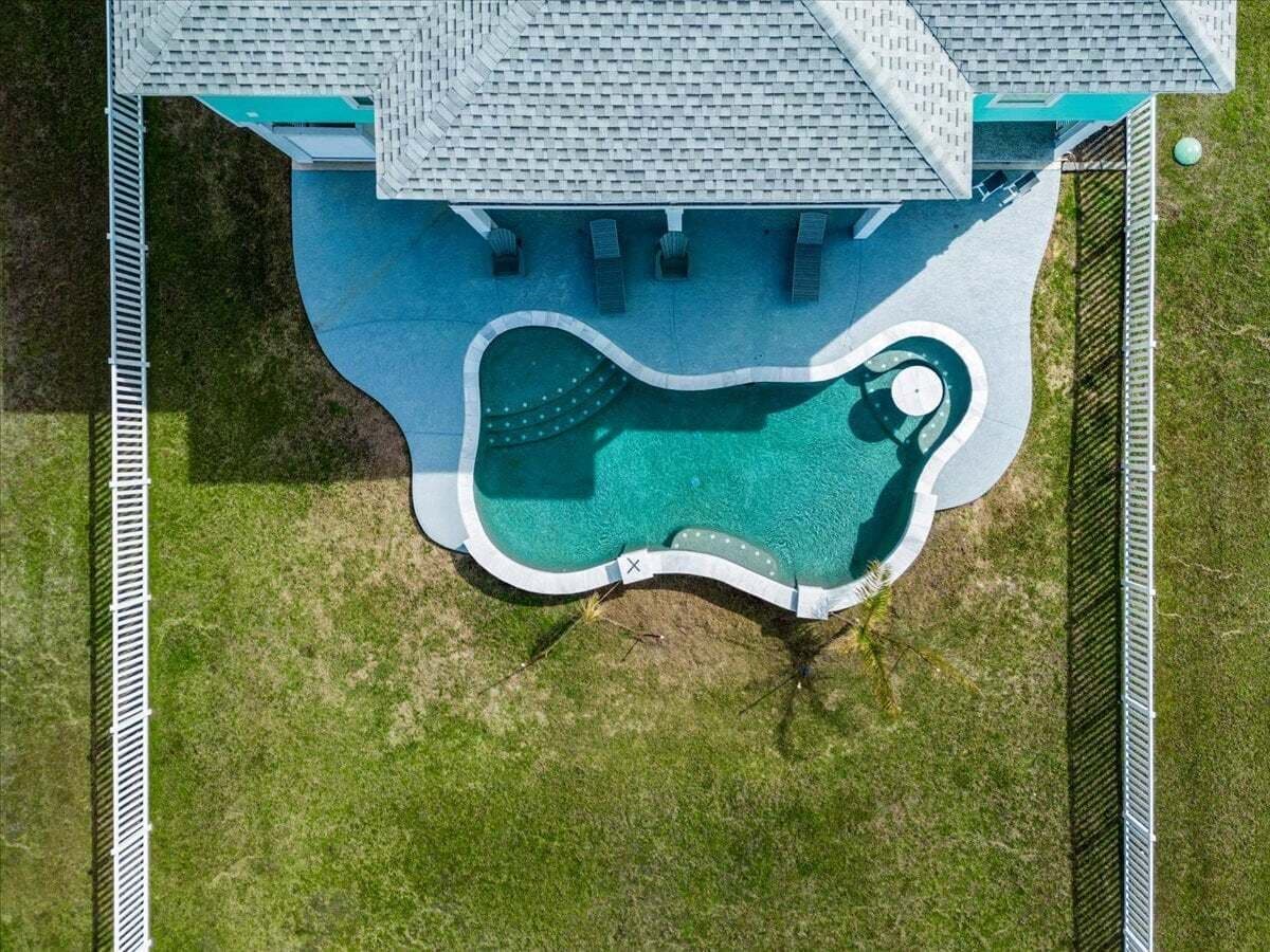 Eine Vogelperspektive zeigt einen Swimmingpool mit ungewöhnlicher Form, umgeben von einem Zaun und einem Gebäude mit geneigtem Dach.