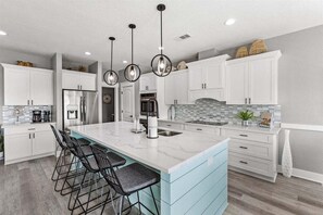 Una cocina moderna con armarios blancos, una isla de mármol, taburetes negros y luminarias colgantes.