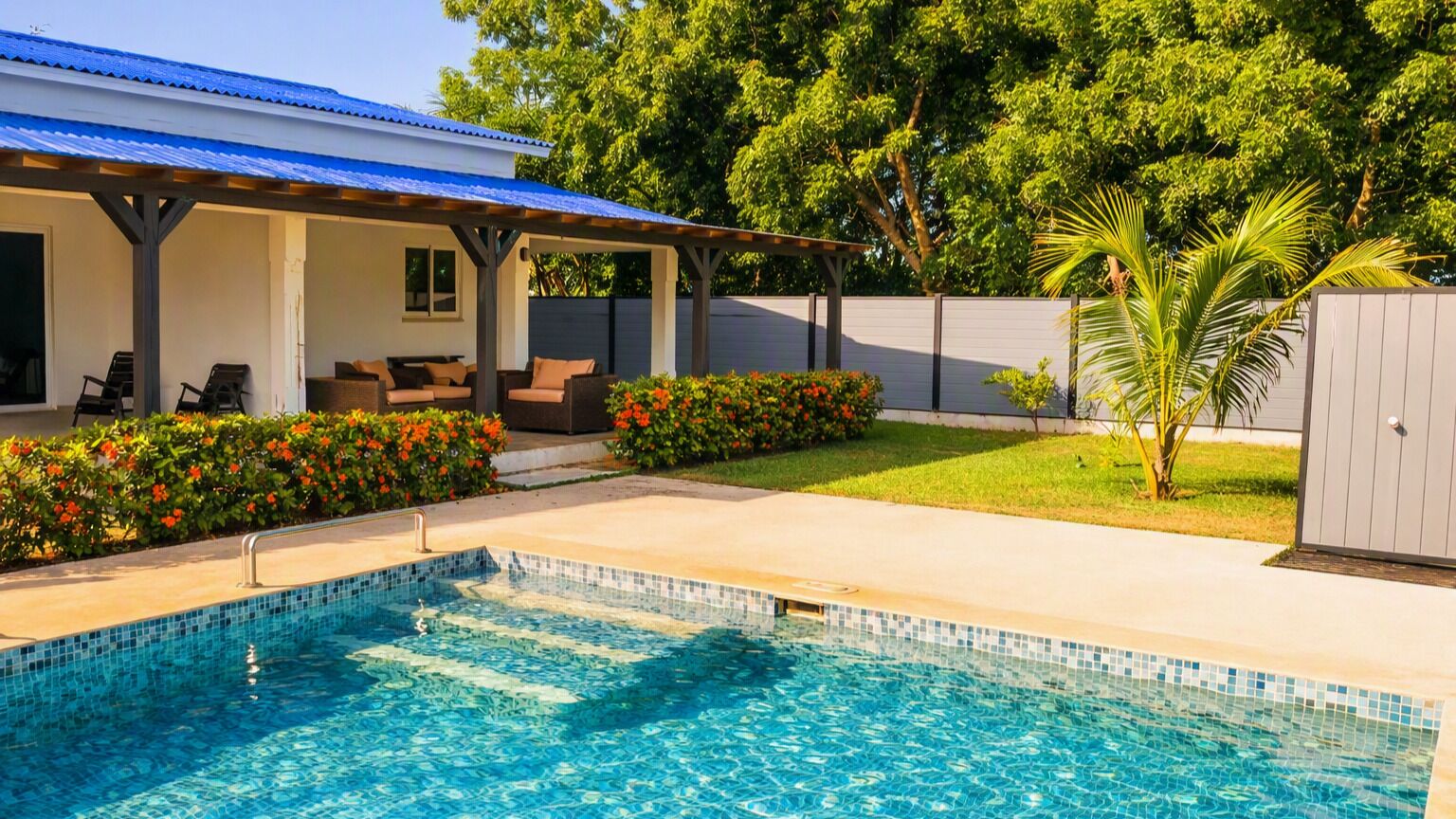 Une piscine avec un bord carrelé, un patio couvert avec des sièges et une cour clôturée agrémentée de plantes fleuries.