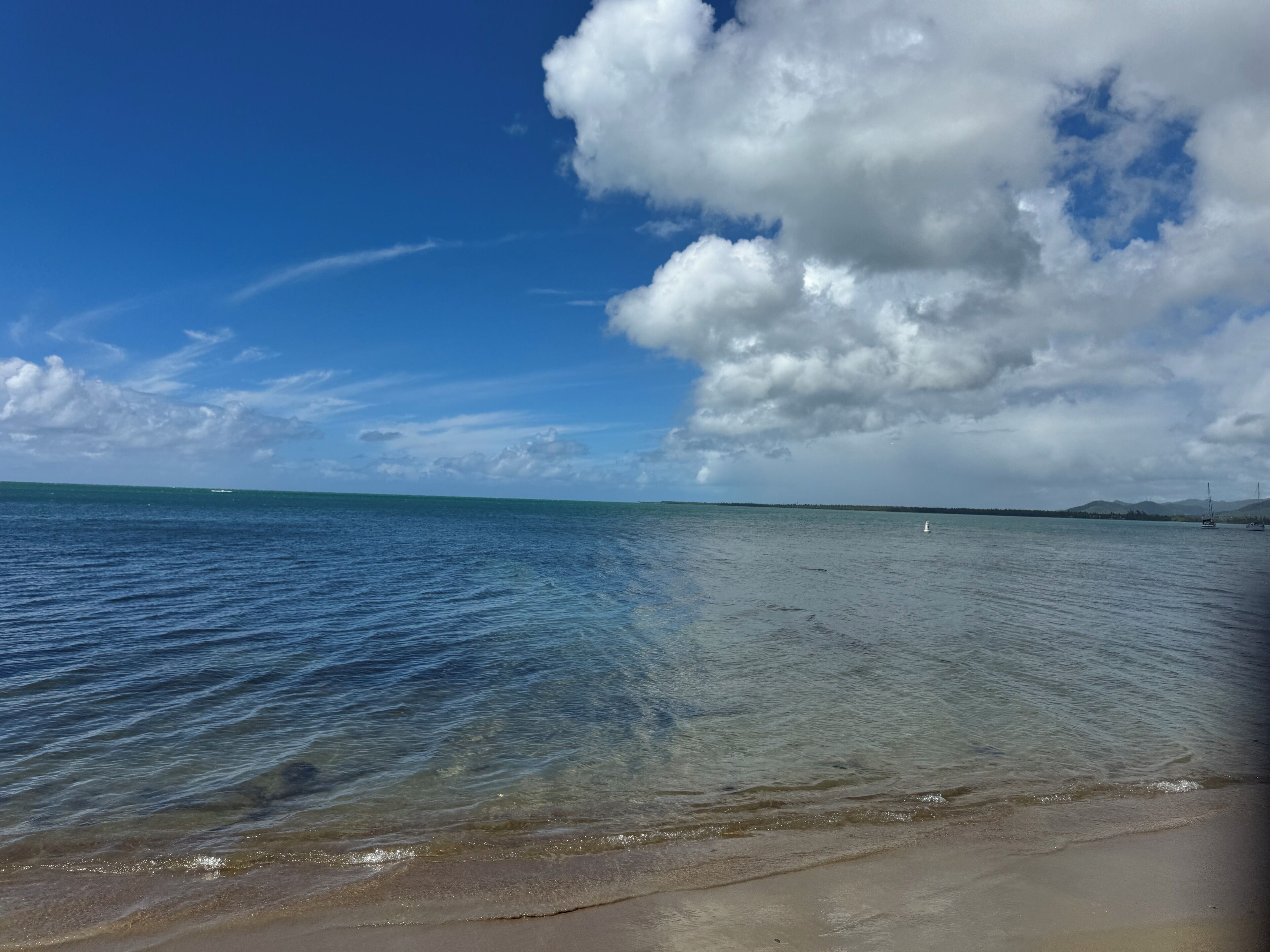 A beach with clear water, a sandy shore, and a distant shoreline with boats.