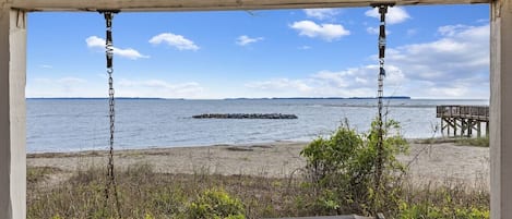 A beach view from a covered area with a bench and a wooden pier in the background.