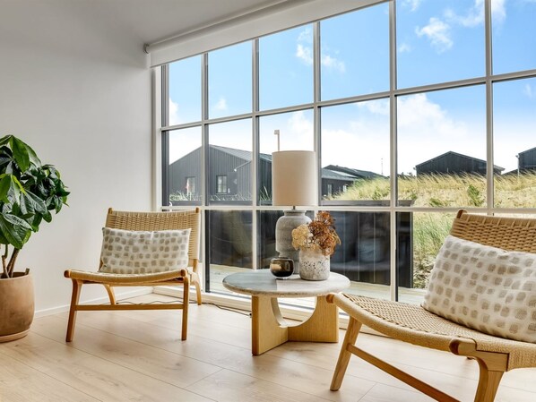 Ein Zimmer mit zwei Korbstühlen, einem runden Tisch, einer Topfpflanze und großen Fenstern mit Blick auf Häuser und Rasen.