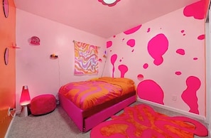 Ein Zimmer mit einem Bett, einer Wand mit abstraktem pinkfarbenem Farbspritzer, einem gemusterten Teppich und einer Lavalampe.