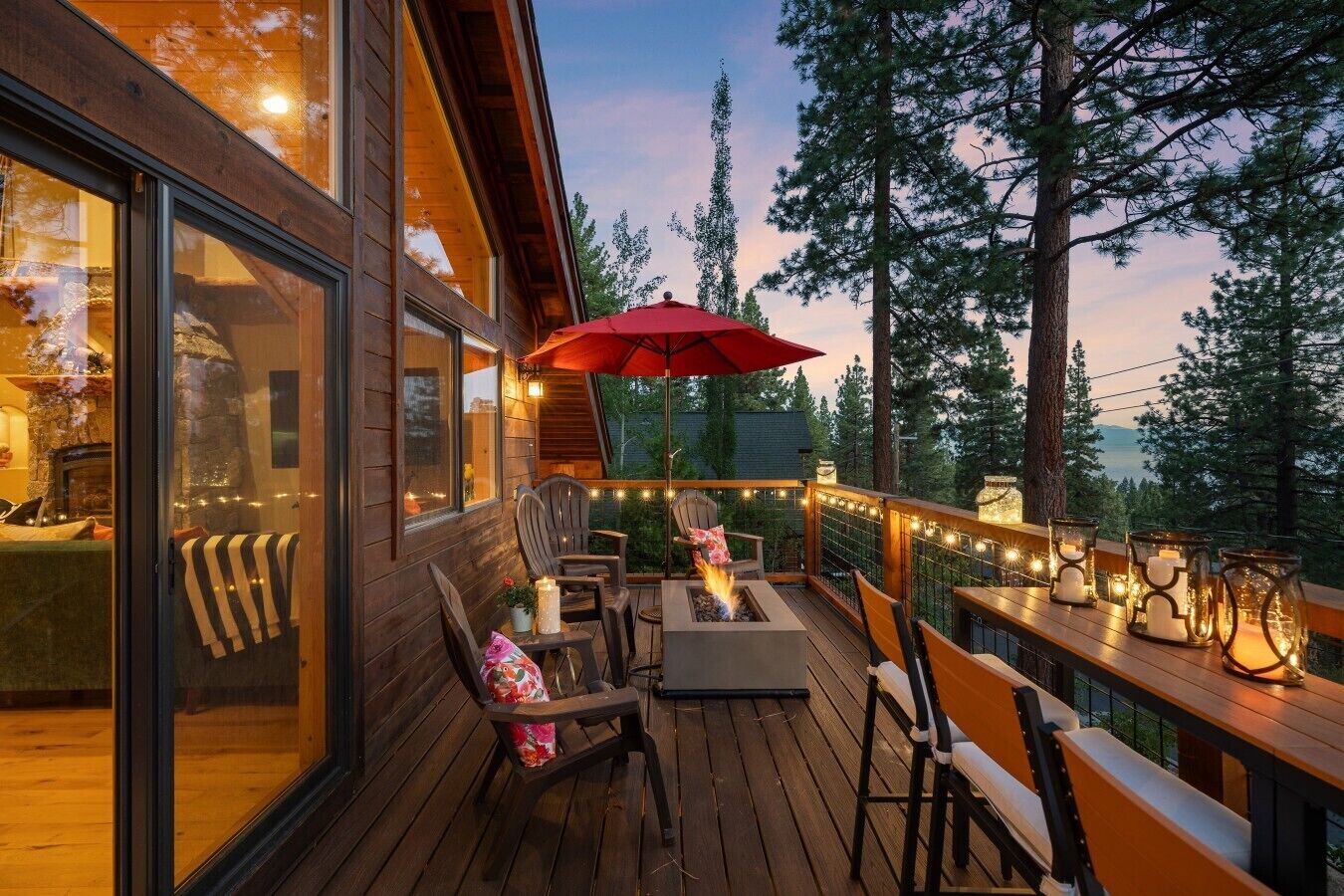A wooden deck with outdoor seating, a fire pit, string lights, and a red umbrella.