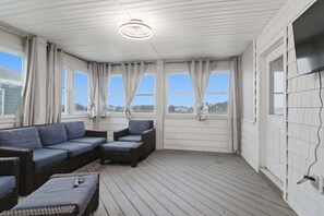 Ein überdachter Außenbereich mit einer Couch und Sesseln, großen Fenstern mit Vorhängen und einer Deckenleuchte.