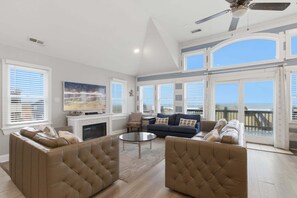 Ein großzügiges Wohnzimmer mit Kamin, bequemen Sitzgelegenheiten und großen Fenstern mit Blick auf den Strand.