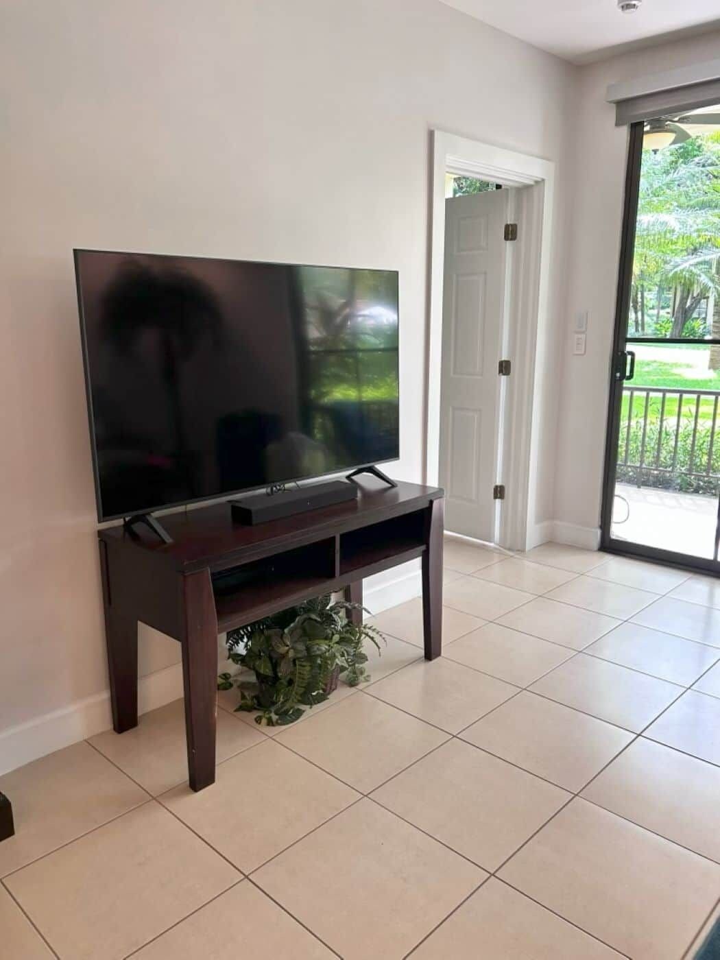 Una sala de estar con televisor de pantalla plana sobre un soporte de madera, piso de baldosas y puerta corrediza de vidrio que da a un balcón.