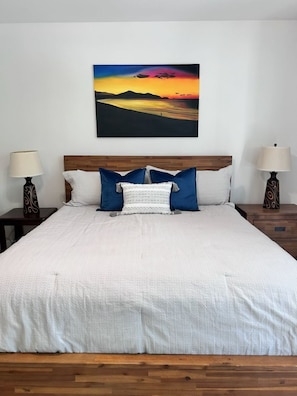 Ein Bett mit weißer Daunendecke, zwei blauen Kissen und einem weißen Kissen, an beiden Seiten zwei Nachttische mit Lampen, vor einer schlichten Wand mit einem Sonnenuntergangsbild.