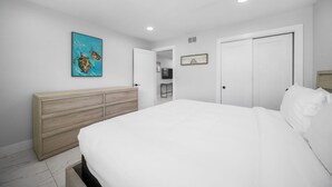 A bedroom with a bed, a wooden dresser, a painting, and a door leading to a bathroom. - 600 #2 Managed by Brampton Park (Deerfield Beach)