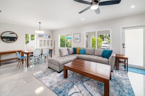 A modern living room with a sectional sofa, a dining area with chairs, a wooden coffee table, a ceiling fan, and large windows with a view of greenery. - 237 Managed by Brampton Park (Delray Beach)