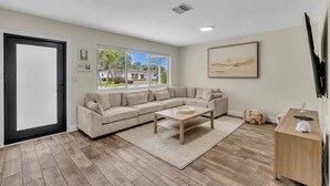 A living room with a sectional sofa, a coffee table, a TV, and a large window with a view of palm trees and houses. - 2240 Managed by Brampton Park (Pompano Beach)