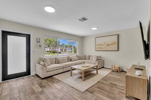 A living room with a sectional sofa, a coffee table, a TV, and a large window with a view of palm trees and houses. - 2240 Managed by Brampton Park (Pompano Beach)