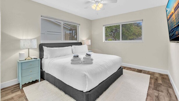 A bedroom with a bed, nightstand, lamp, ceiling fan, windows, and a TV. - 2240 Managed by Brampton Park (Pompano Beach)