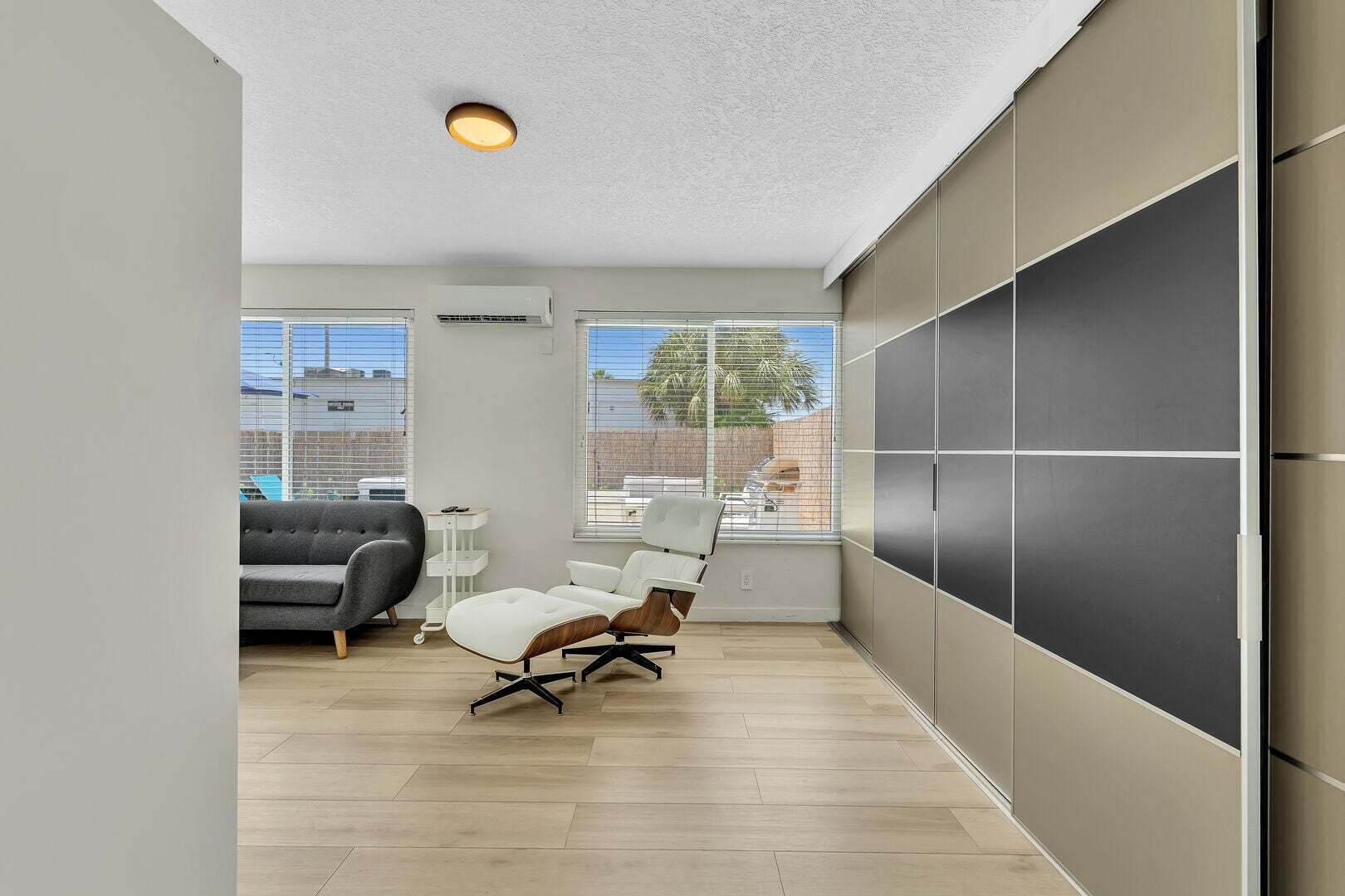Un salon moderne avec un canapé, un fauteuil blanc, un mur sur lequel est fixé un téléviseur à écran plat et un luminaire au plafond.