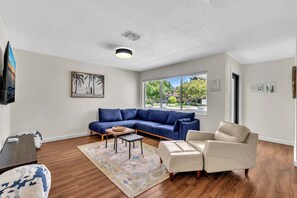 Ein Wohnzimmer mit einem blauen Sofa, einem beigen Sessel, einem Couchtisch und einem großen Fenster mit Blick ins Freie.