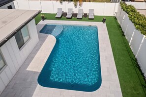 Ein rechteckiger Pool mit abgerundeter Kante, umgeben von einem weißen Zaun und einer kleinen Rasenfläche mit Liegestühlen.