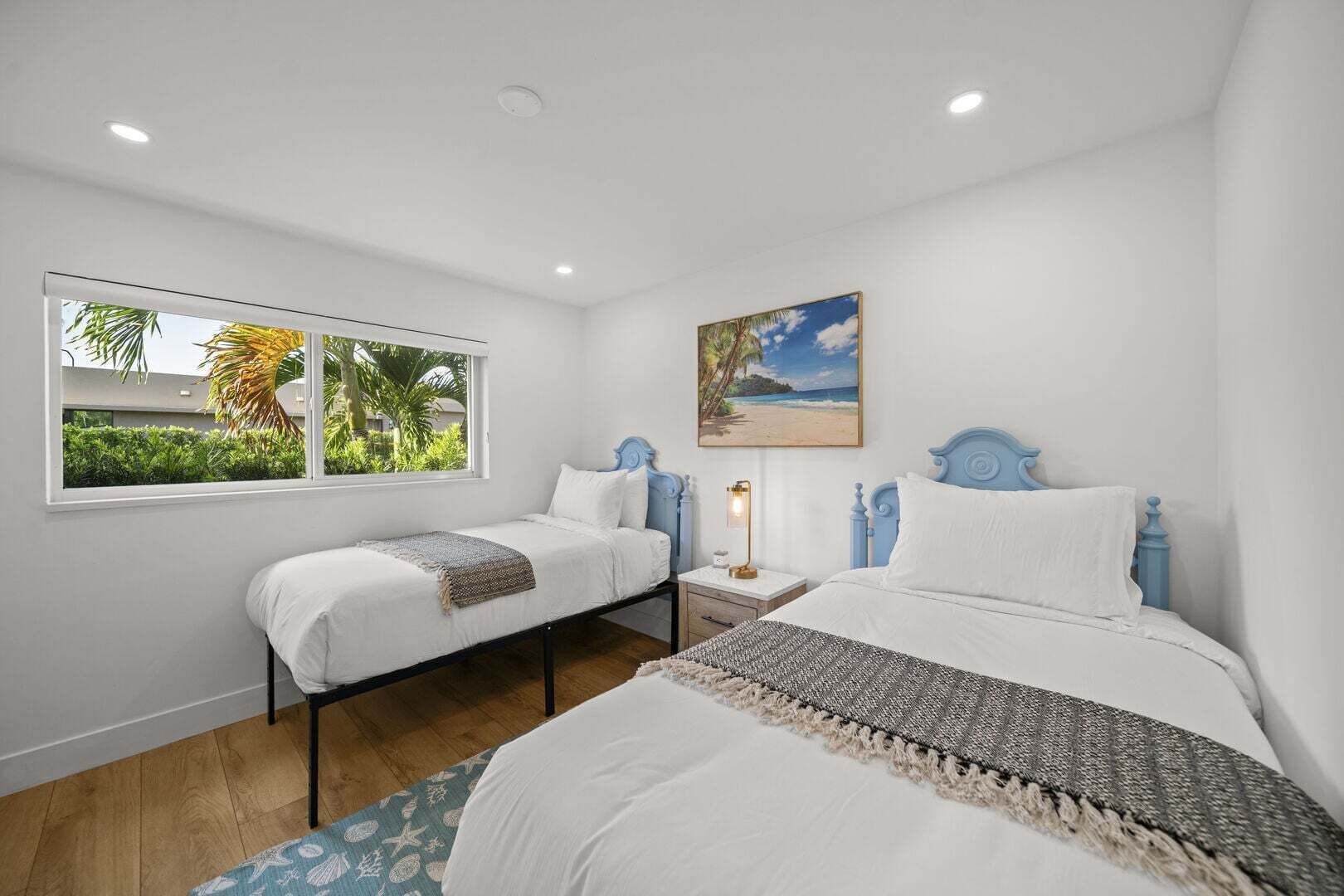 Une chambre avec deux lits, une fenêtre donnant sur un paysage verdoyant, un tableau représentant une plage et une table de chevet avec une lampe.