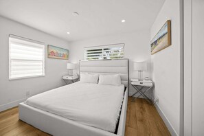 Ein Schlafzimmer mit einem Bett, zwei Nachttischen, einem Fenster mit Jalousien, einem an der Wand befestigten Regal mit Fotos und einem Gemälde.